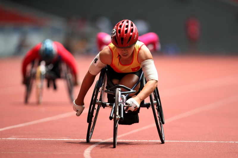 PU跑道上，一位身穿黃色無袖運動裝、黑色短褲、頭戴紅色單車安全帽的女選手，坐著三輪手搖車，壓低身子衝刺中，身後有其他手搖車選手，正在進行輪椅競速。